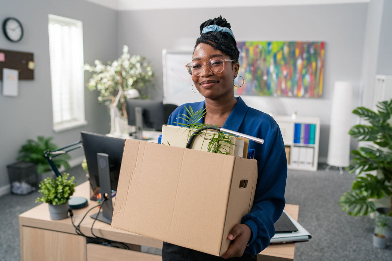 Stylish businesswoman wearing elegant blue clothes and glasses is promoted to managerial position changes office, leaves company, carries carton with packed accessories, change of job