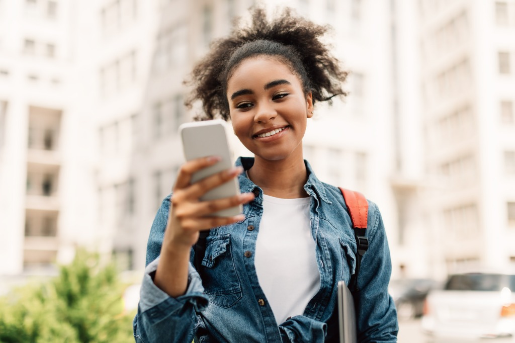 cheerful african american student using mobile phone standing outside